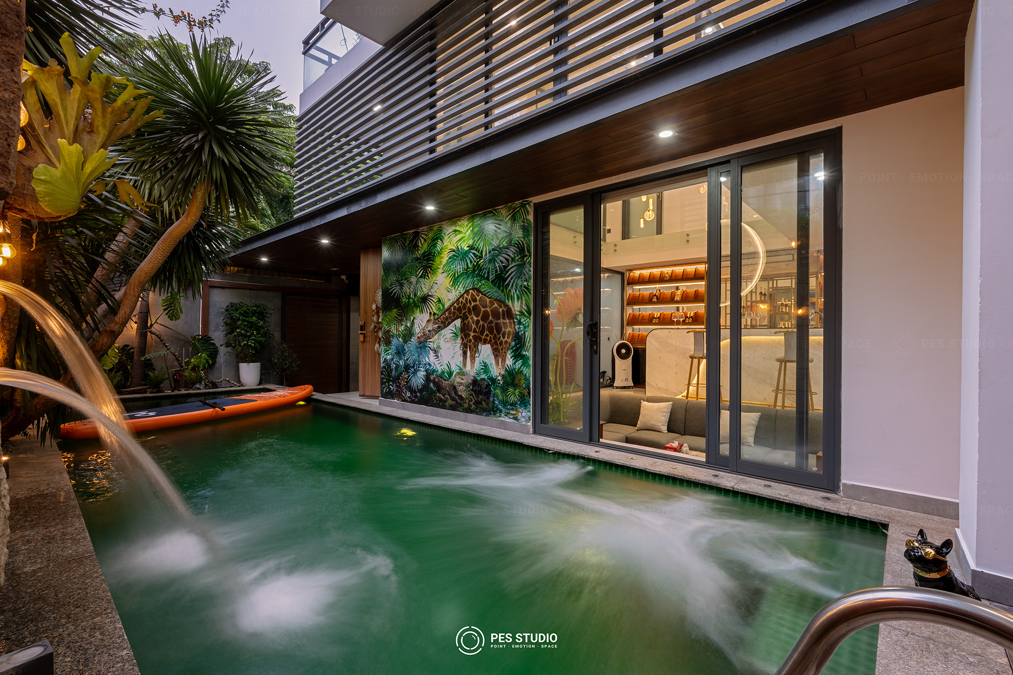 Hồ bơi villa nhìn qua vách kính lớn với tường cây và đèn hắt ấm